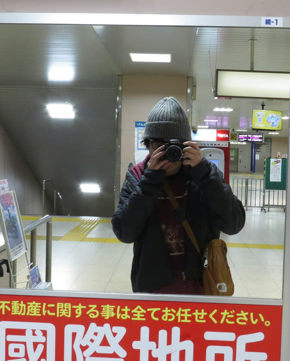 A mirror in a Japanese railway station, reflecting a person in a grey knitted cap holding a camera up to their face.
